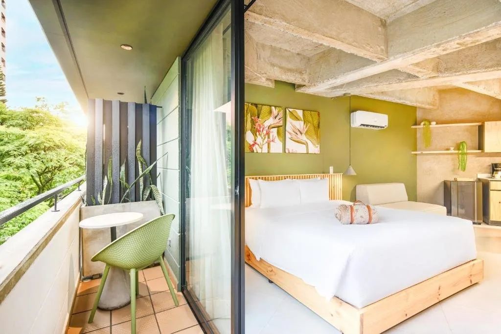 Habitación con cama, techo de concreto a la vista y balcón con mesa y silla verde